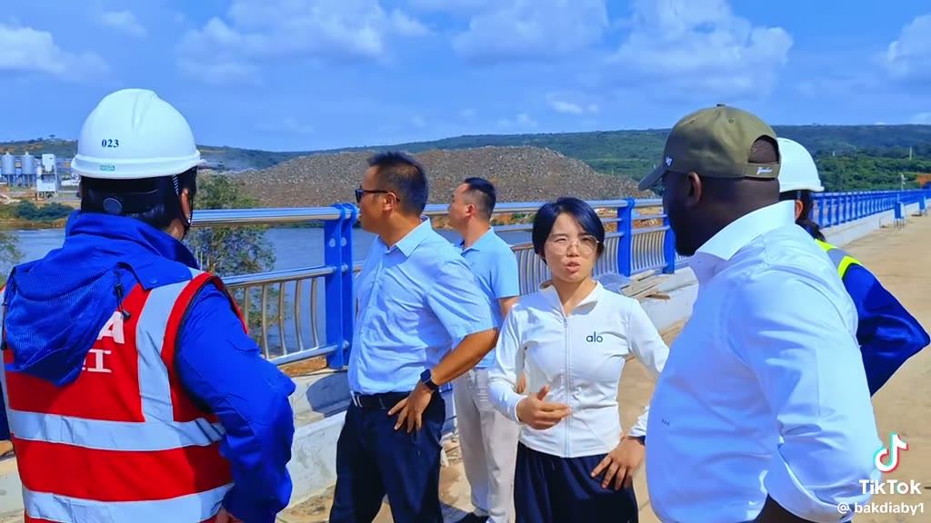 Visite d'un grand ouvrage en béton : un groupe en équipements de protection sur le tablier de l'ouvrage, avec plan d'eau et silos industriels en arrière-plan