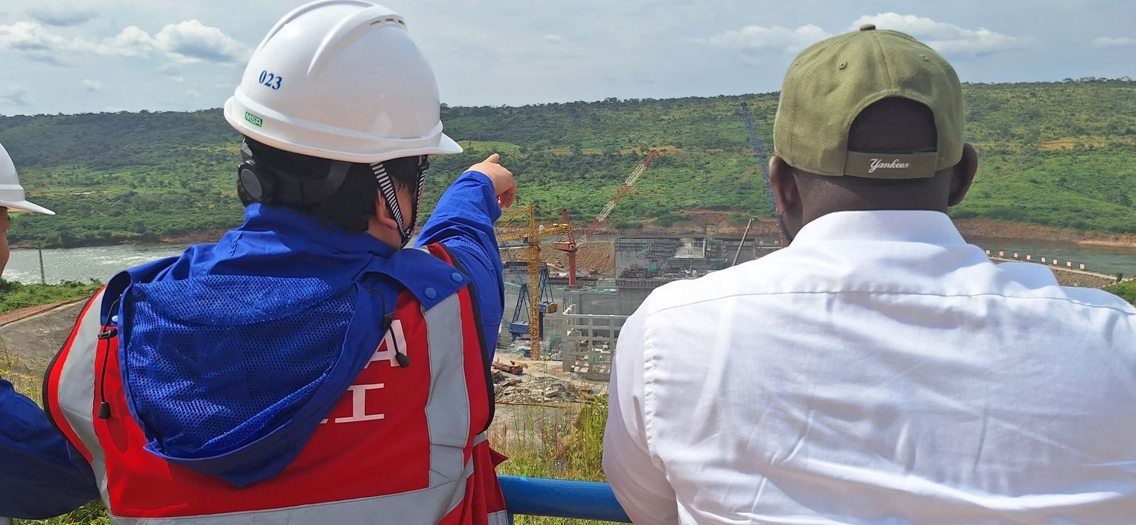 Visite de chantier : un ingénieur partenaire désigne un barrage hydroélectrique en construction — grues, ouvrages en béton et fleuve — aux côtés du Président-Directeur Général